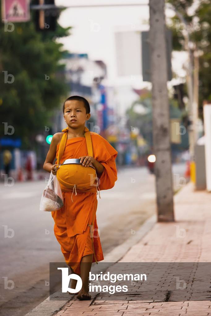 Thailand, Chiang Mai, Young boy wearing orange robe walking down edge of street (photo)