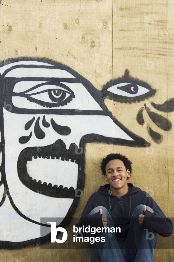 UK, Teenage boy sitting in front of graffiti wall of cubist style face, Hastings (photo)