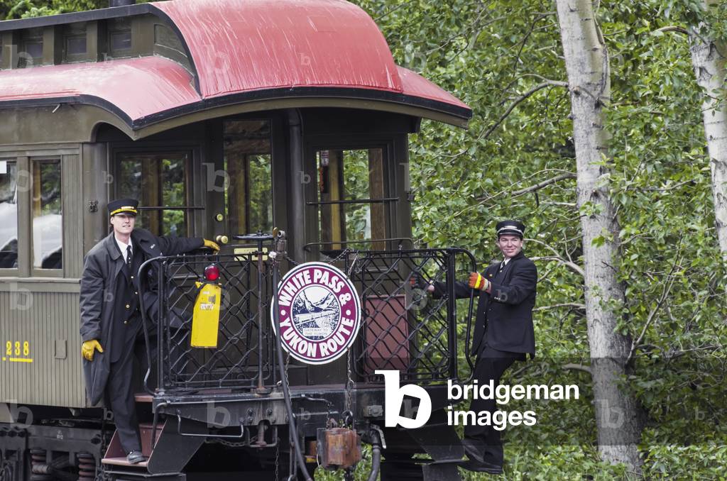 Two conductors watch for pedestrians crossing tracks as White Pass & Yukon Route RR train is backed up behind downtown depot; downtown historic district Skagway Alaska (photo)
