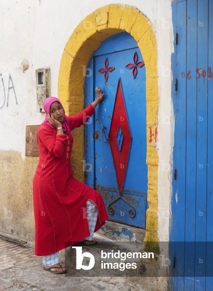 A woman standing at the colourful door of a house talking on her cell phone, Essaouira, Morocco (photo)