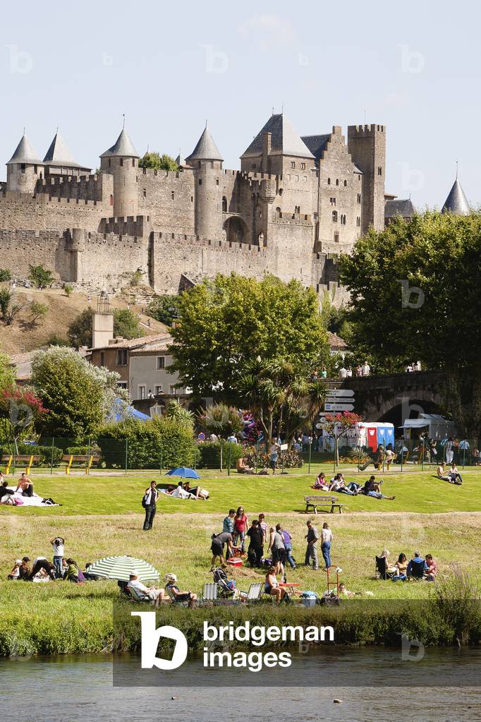 People gathering on the lawn by the river with castle and ramparts in the background, Carcassonne, Languedoc-Rousillion, France (photo)