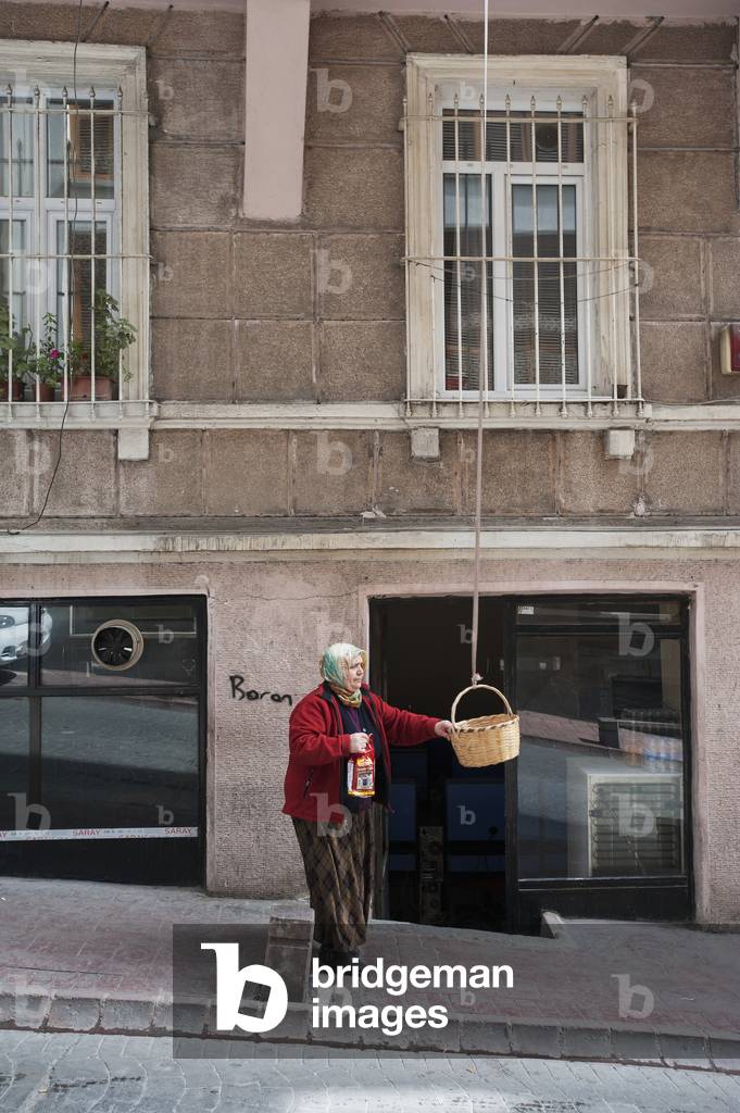 A woman stands holding a basket which hangs from an upper level of a building, Istanbul, Turkey (photo)
