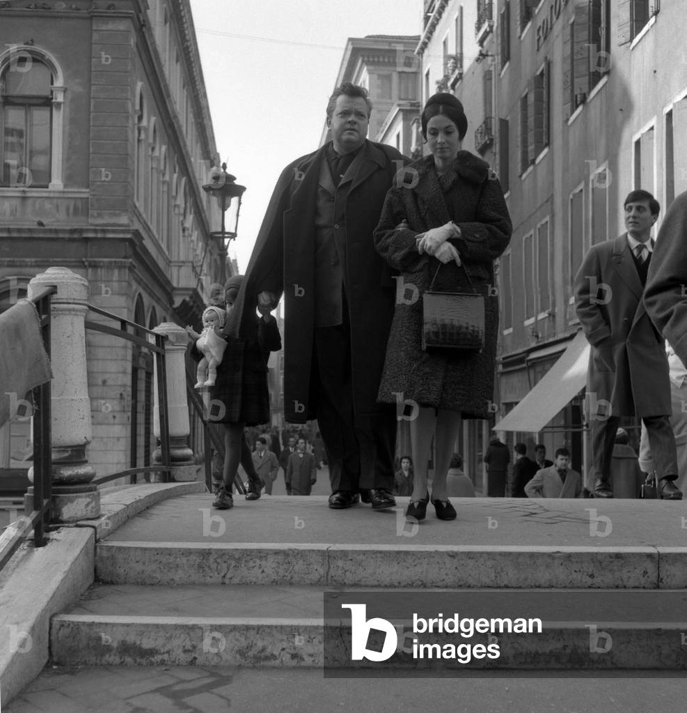 American film director Orson Welles, wearing a coat and a scarf, walking on a bridge with his wife Paola Mori, her hair in a bun, holding a crocodile bag, next to their daughter holding a doll, Venice, 1956