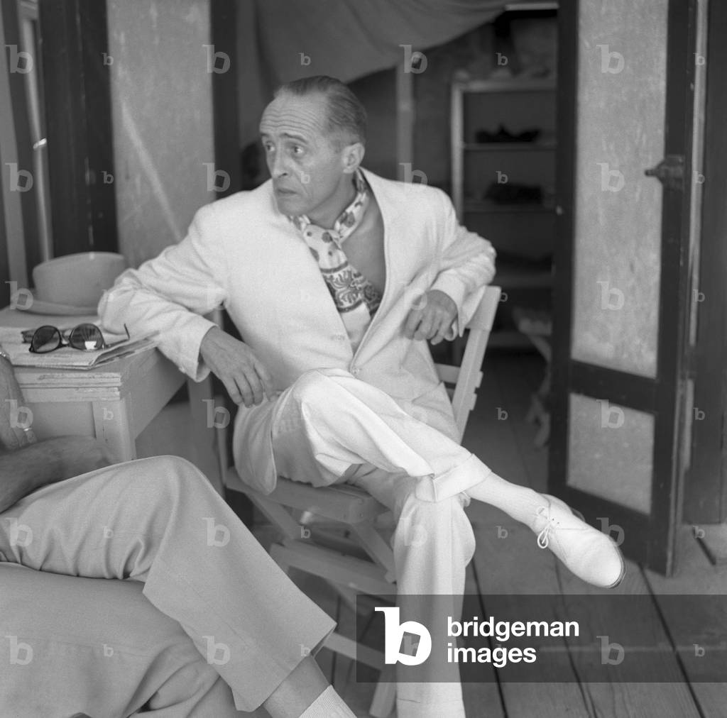 French director Rene' Clair, wearing a white suit and a foulard, portrayed while sitting on a chair and leaning on a table, Venice, 1958