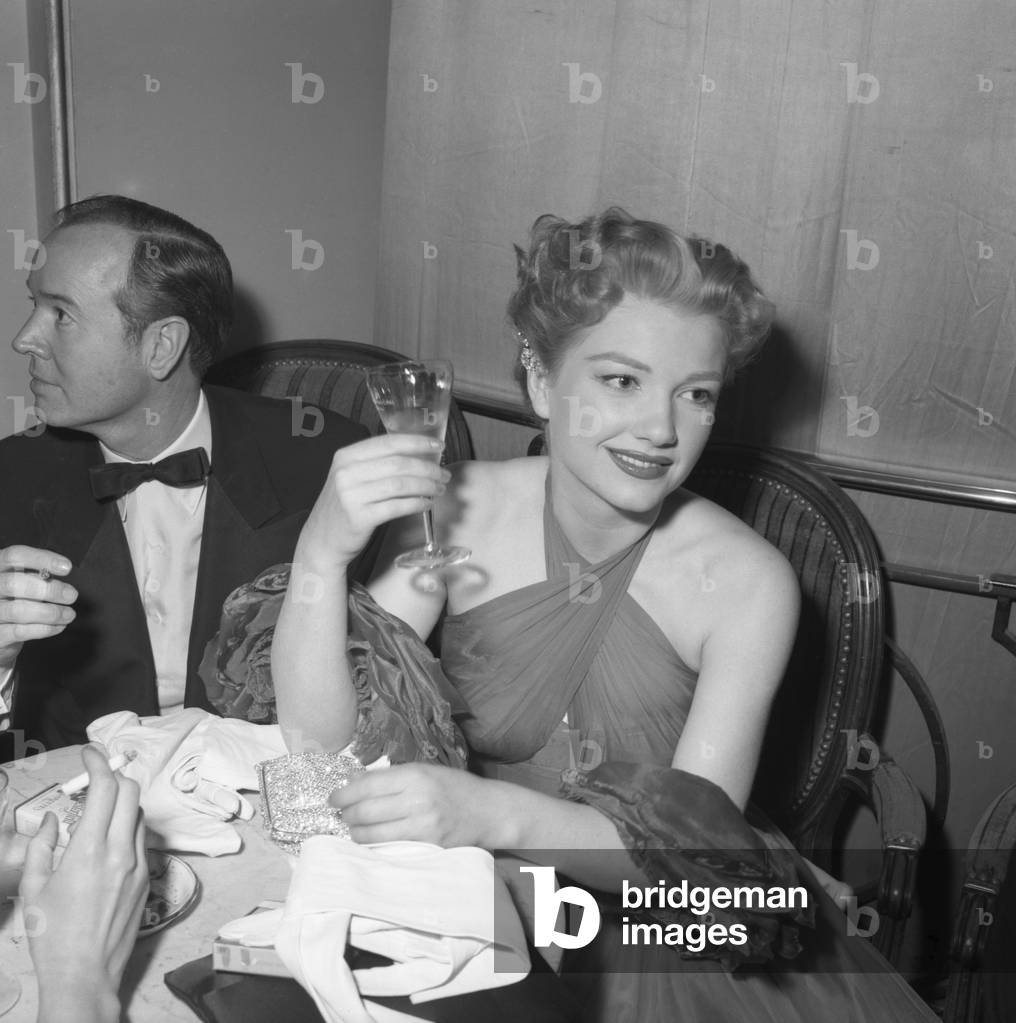 American actress Anne Baxter, wearing an evening dress and holding a drink, sitting at a table with a man wearing a tuxedo and a bow tie, Cannes, 1953