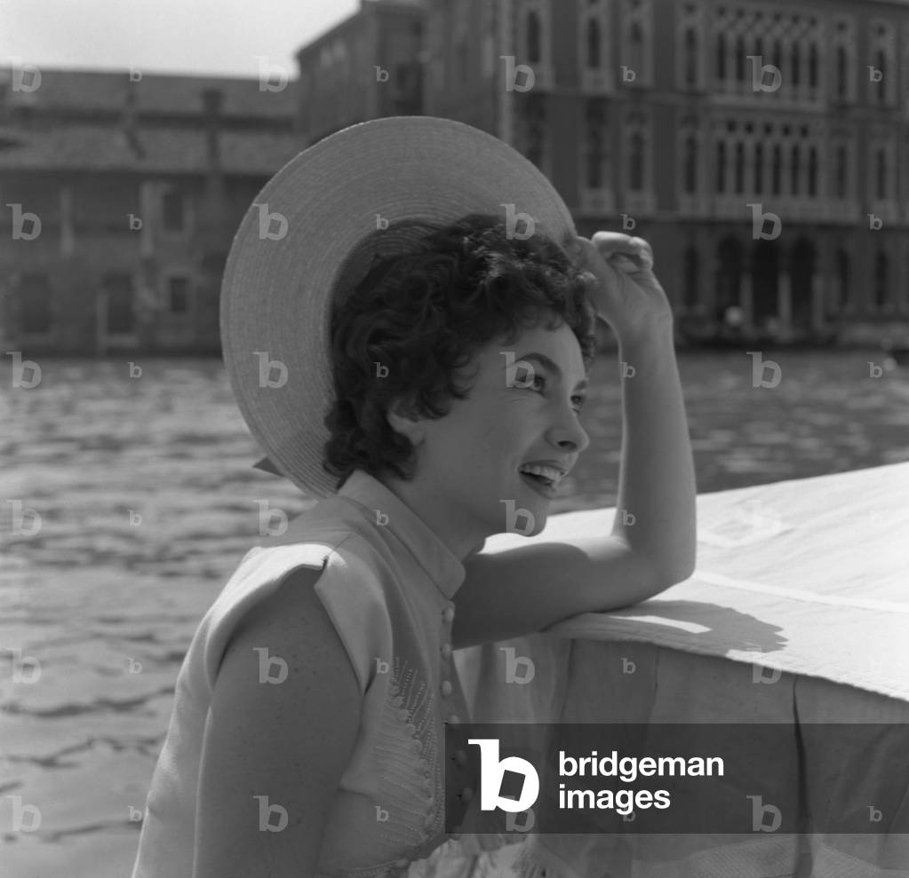 Italian actress Gina Lollobrigida wearing an embroidered dress and a straw hat, portrayed from the side, standing on a water taxi in The Canal Grande, Venice, 1954