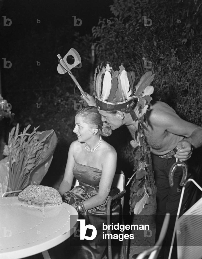American actress Joan Fontaine, wearing an evening dress and a necklace, her handbag on the table in front of her, a man wearing indian-american native disguise and holding a toy axe, during the Pecos Bill Party, Venice, 1952.