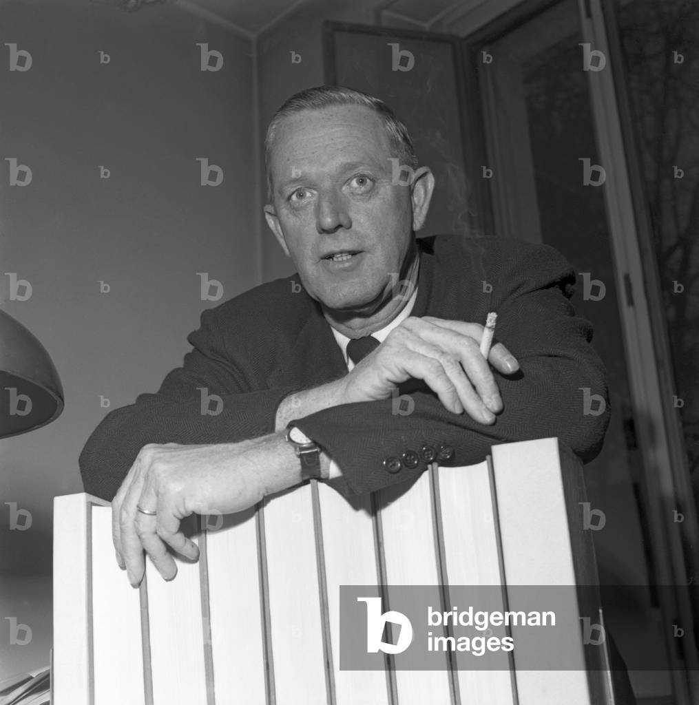 American author Erskine Caldwell, wearing a suit and a tie, smoking a cigarette portrayed while leaning on some books in his studio in Milan, 1953