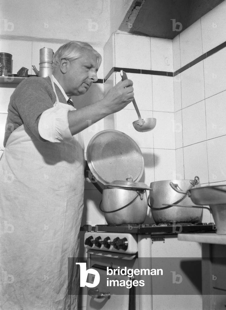 Italian painter Giorgio De Chirico in his kitchen, wearing an apron, opening a pot and holding a dipper, Venic