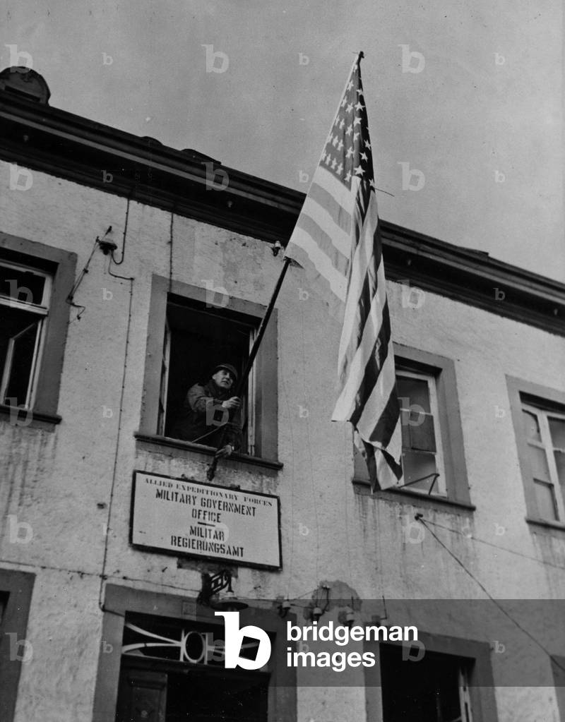 WWII:Daleiden (Germany) 24 February 1945: A corporal of the 6th DB US deploys the Star Banner on the facade of the Office of the Allie Military Government.