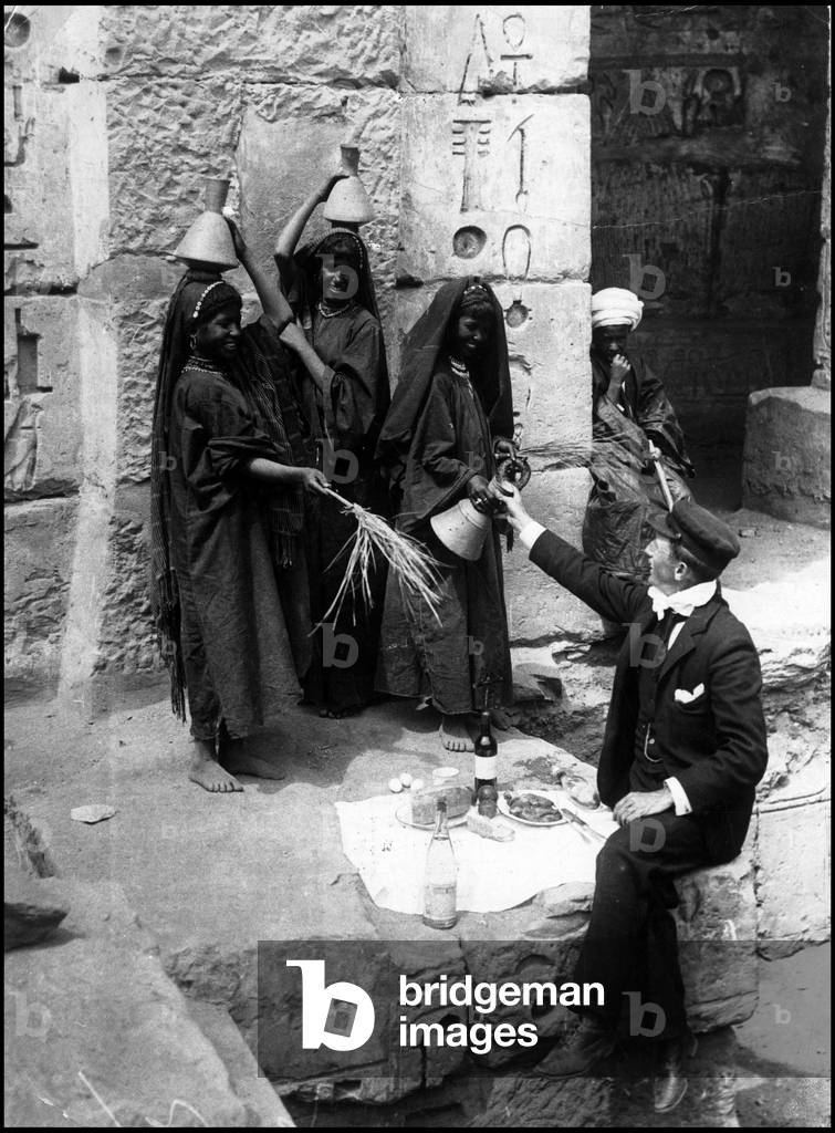 Upper Egypt around 1900: A young tourist in the Temple of Medinet Habu of Pharaoh Ramses III. Young girl gives him a jug of water