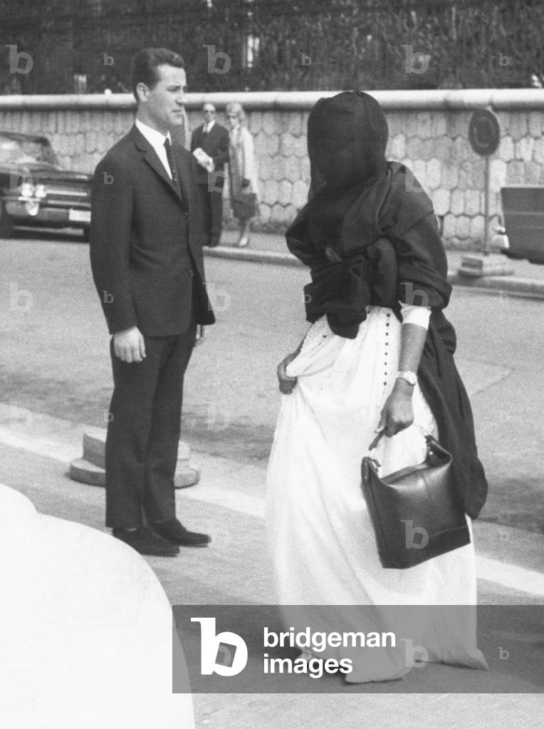 Wearing a dark veil, one of the wives of King Saud of Saudi Arabia enters the Hotel Negresco. King Saud was on a recuperative vacation in Nice, France. March 28, 1963. A private detective stands near