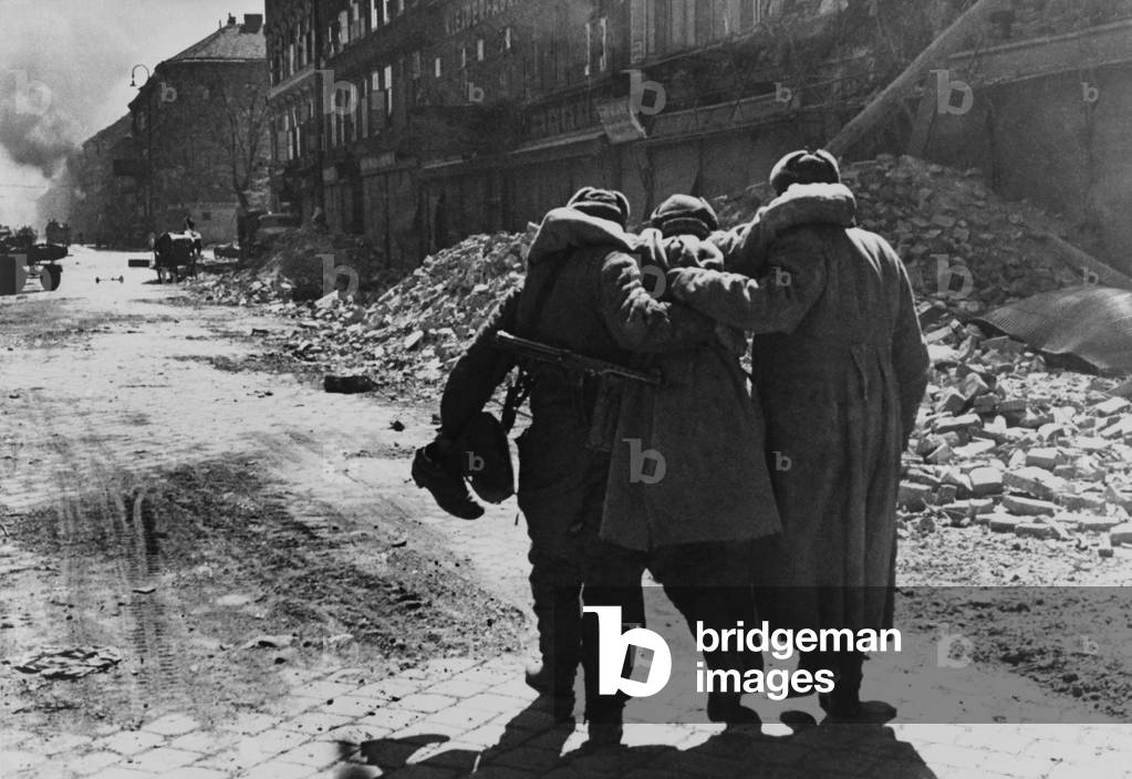 Wounded Soviet (Russian) soldier helped by two comrades in a street filled with rubble, Vienna. 1945 World War 2 photography by Evgenii Khaldei