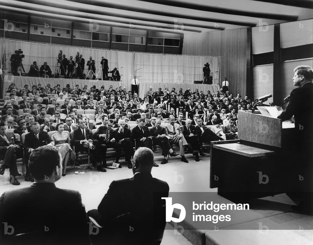 President John Kennedy speaks to reporters during a press conference. Press Secretary Pierre Salinger and Associate Press Secretary Andrew Hatcher sit near the podium at State Department Auditorium, Washington, DC. 1961-63