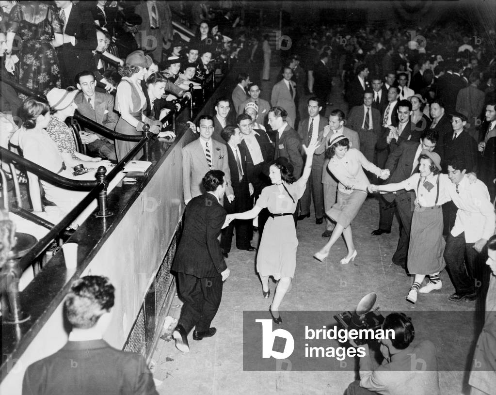 Couples jitterbug dancing on a dance floor, with a photographer in the foreground and the audience in raised seating on the left, photograph by Alan Fisher, 1938