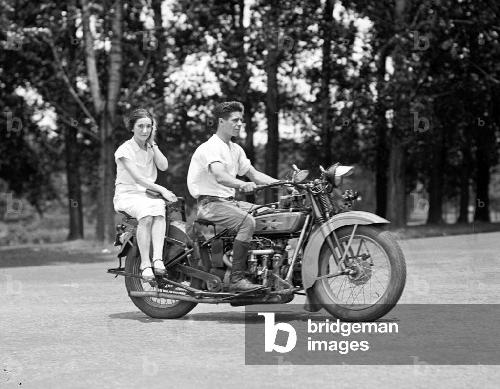 A young man drives a motorcycle while a young woman rides side saddle behind him. c. 1929-1930