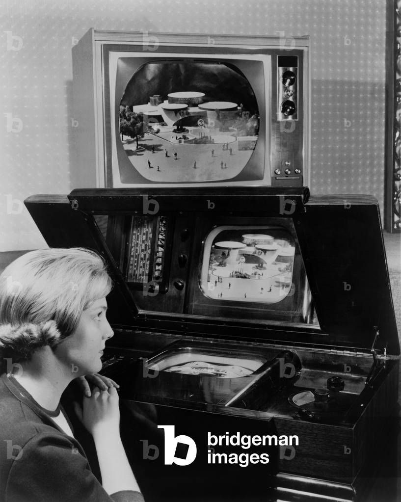 Woman looking at two television sets. One, similar to a set demonstrated at the 1939 New York World's fair, has a small screen incased horizontally in a large wooden cabinet and is viewed from a reflective mirror. The other, now displayed 25 years later at the 1964 New York World's fair, is a large screened color television