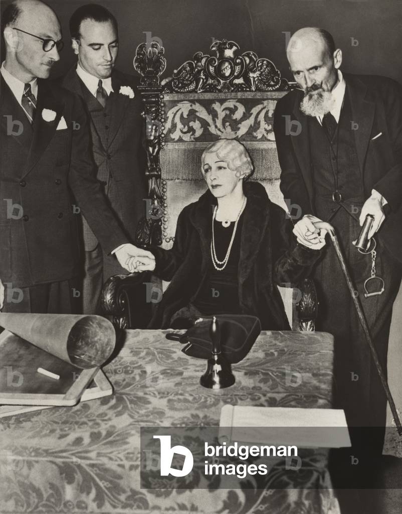 Mrs. Bess Houdini, widow of the great magician, sitting in a chair with her business manager, Edward Saint (to her left holding handcuffs), and others, during a seance, in an effort to contact the spirit of Harry Houdini. 1936