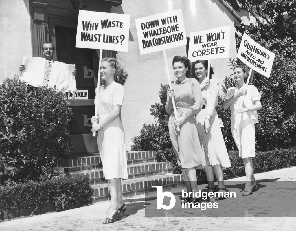 The Lane Sisters with Gail Page protest famous Hollywood costume designer, Orry-Kelly. Aug. 28, 1939. The Warner Brothers, designer Orry Kelly holds a corset, the actresses picket with signs reading: We Won't Wear Corsets, Down with a Whalebone Boa Constrictor, and Our Figures Need No I improvement