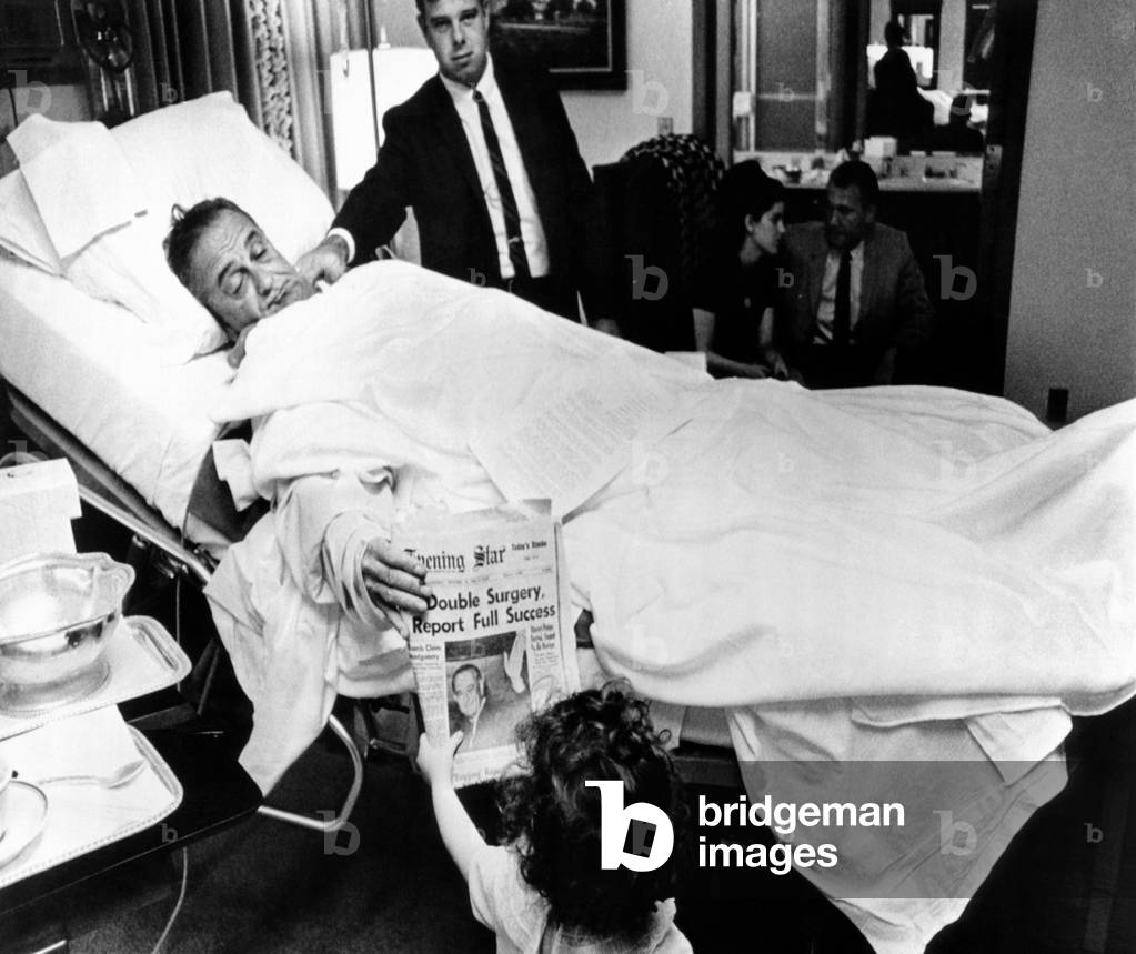 President Lyndon Johnson after gall bladder surgery. He shows a headline about his operation to Courtenay Valenti, daughter, Jack Valenti, at Bethesda Naval Hospital. In the background are Pat and Luci Nugent. Oct. XX, 1965