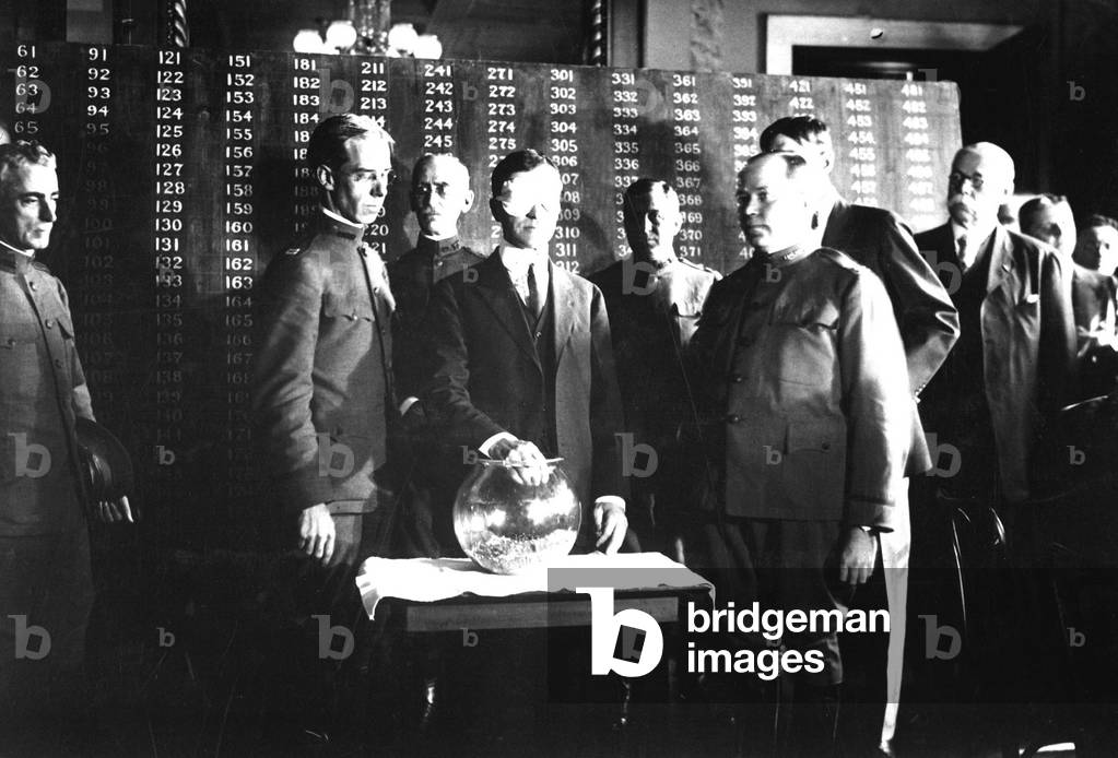 Secretary of War Newton D. Baker, wearing a blindfold, initiates the second draft of World War I. He draws the first number, 246, out of a glass bowl to start the draft. Behind him was a black board of numbers that mapped to birthdates and draft classifications. June 1918