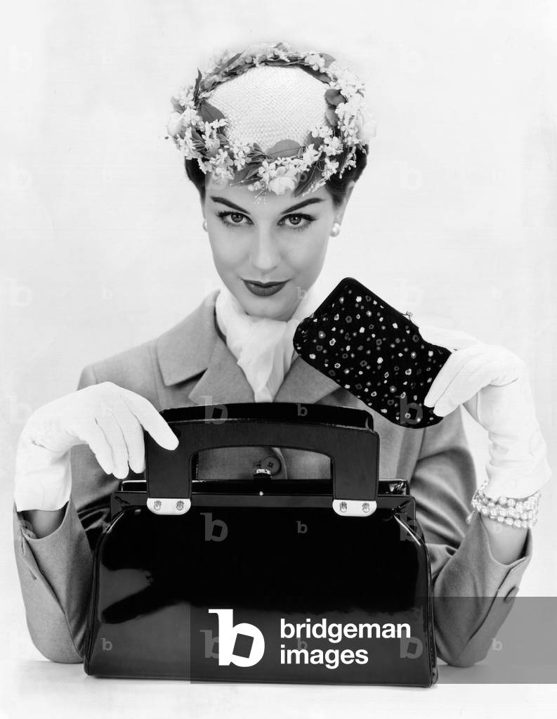 Women's accessories include a square cut, top opening, black patent leather purse. A small forward worn hat with floral edging and gloves add to the crisp formality of the look. She wears a tailored jacket and defining eye and lip makeup. March 1956