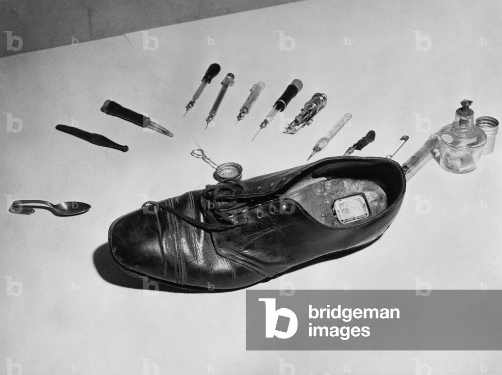 Law enforcement photo of a shoe with metal box in cut out section of heel used to smuggle drugs into a detention facility. For dramatic effect the shoe is surrounded by drug paraphernalia. 1955