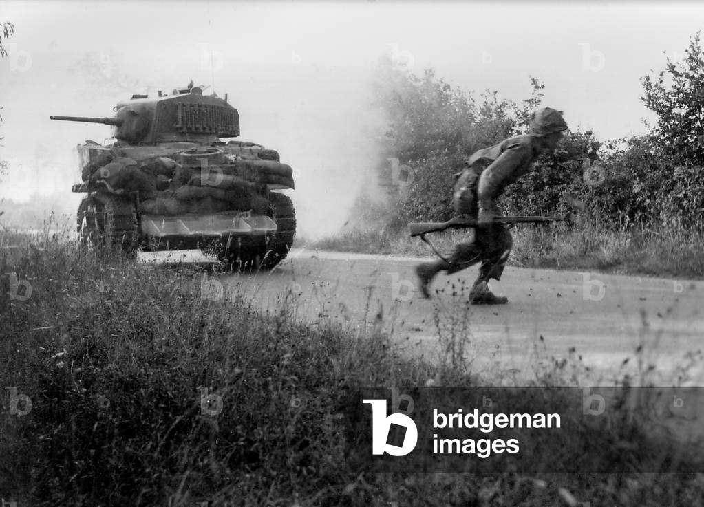 U.S. soldier crosses a road cautiously under the cover of a light tank in the Hedgerows of Normandy. The tank is equipped with the hedgecutter device developed in July 1944. Normandy Campaign, France, World War 2