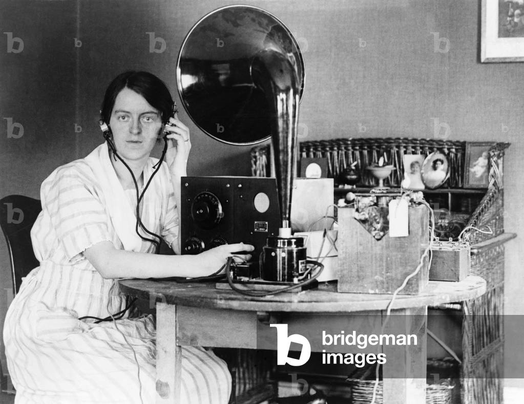 Woman listening to radio in her home receiving a personal radio message telling her when her husband will arrive home. 1922