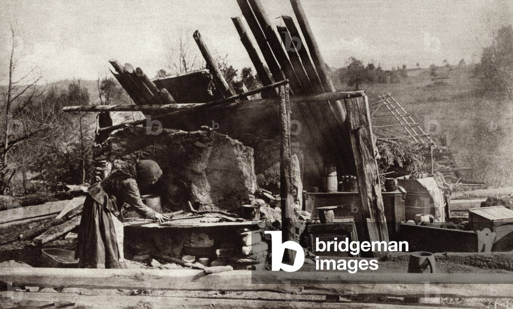 World War 1. Russian peasant woman in her battle ruined home on the Eastern Front. She placed her baby in the cradle at right and is attempting to prepare a meal. c. 1915