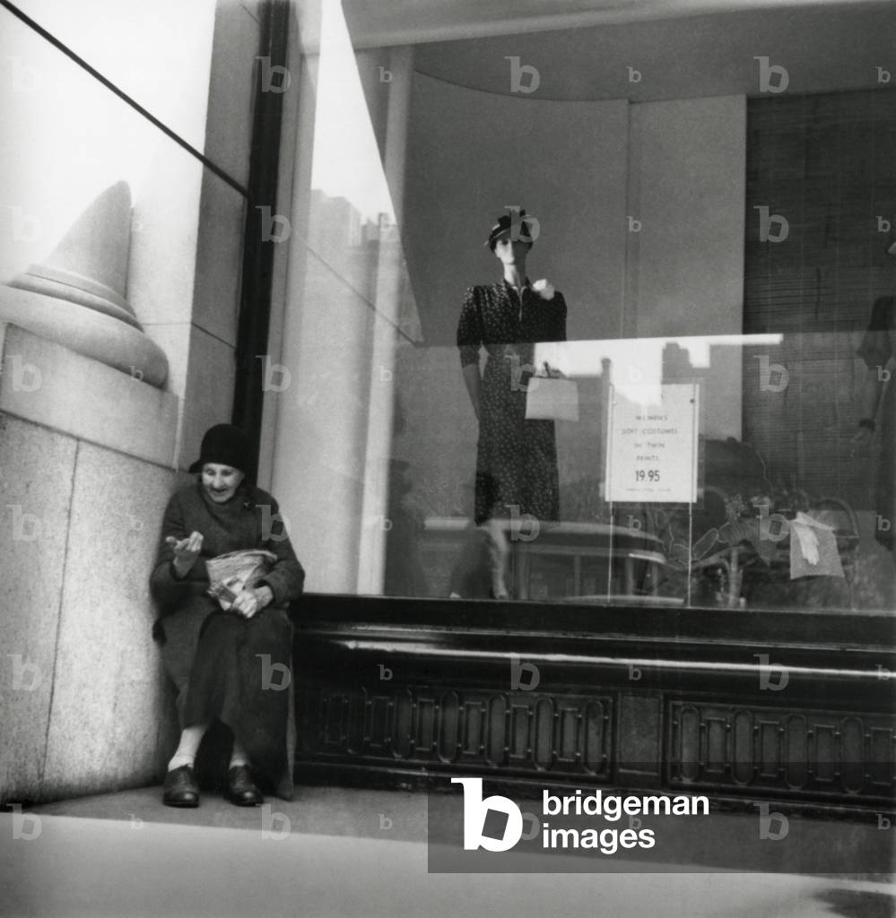 Elderly woman beggar selling pencils during the Great Depression, 1938. She is sheltered against an urban dress shop window. The photo was taken New Deal photographer and featured in 'One Third of the Nation', by Arnold Eagle and David Robbins