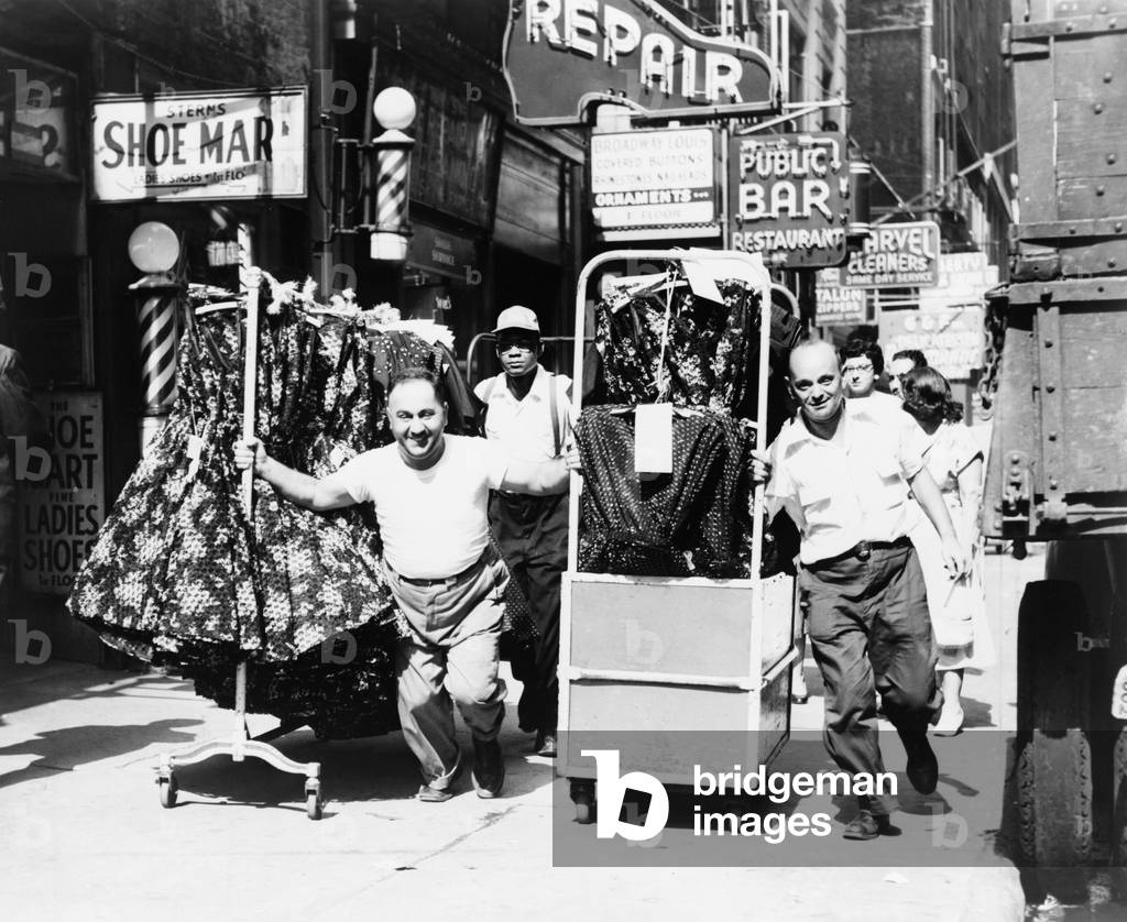 Men pulling racks of clothing on busy sidewalk in Garment District, express the vitality of mid-20th century New York. 1955