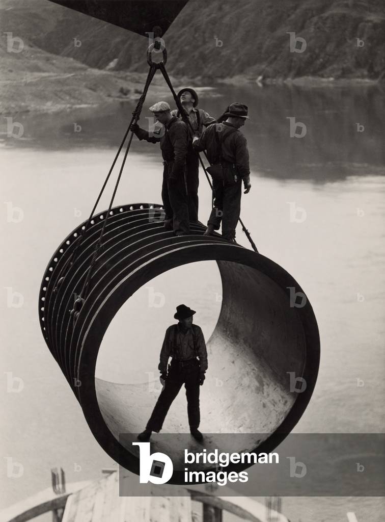 Four workers riding on a large casing section of pipe suspended from cable as it is moved into position during construction (1933-42) of the Grand Coulee Dam, Washington