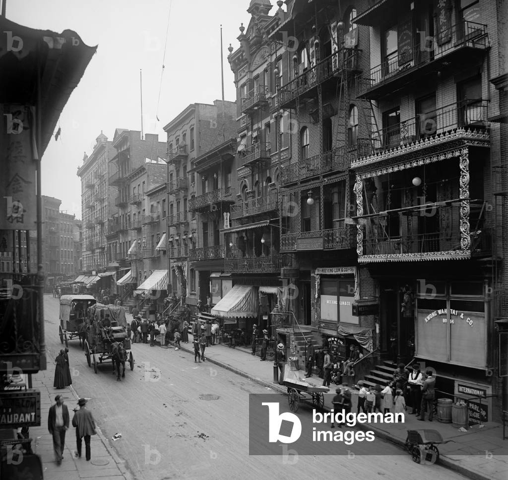 Mott Street in New York City's Chinatown presents a cosmopolitan scene mixing Asian and American-European dress and faces. The street is lined with restaurants, laundries, and pushcarts. c. 1900