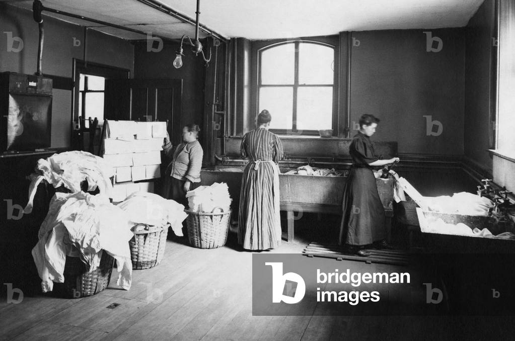 Laundry with women working at large sinks, one operating a hand cranked wringer, and another at a drying rack placed near a stove. Boston, Massachusetts, 1905