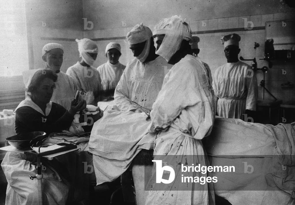 Masked surgeons perform an operation while a nurse administers ether to anesthetize patient. c. 1920