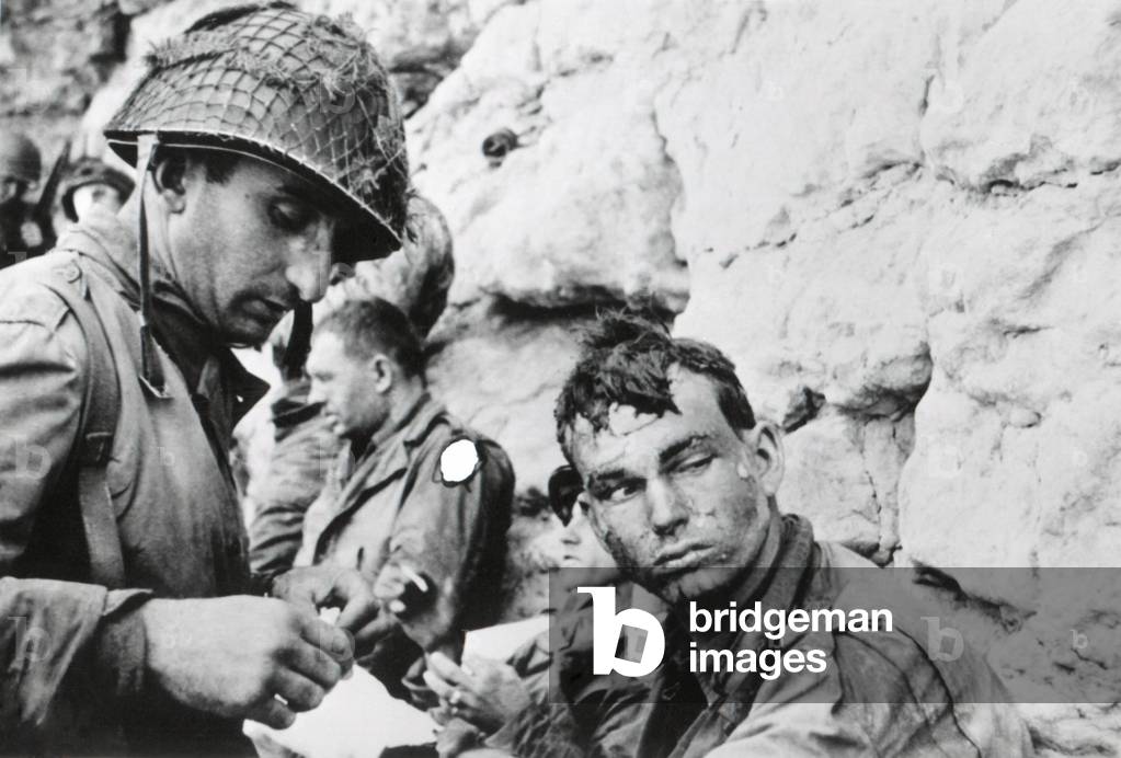 U.S. soldier who was injured during landing on Normandy beachhead, on D-Day plus 1. A medic bandages his hand. The marks on his face came from water spray while landing. June 8, 1944. World War 2