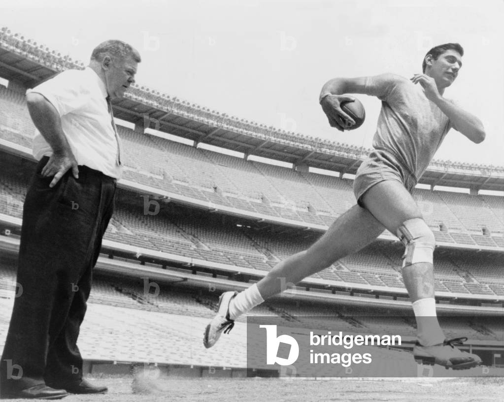 Joe Namath (b. 1943), quarterback with the New York Jets, running during light workout to test his injured knee, as coach Weeb Eubank observes. After his football career, Namath had knee replacement surgery. 1965