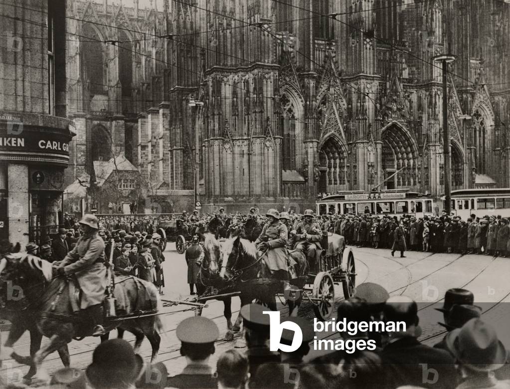 German troops entering Cologne, March 17, 1936. Civilians watch a parade of German officers, some on foot, others on horses pulling carts holding officers. By entering the Rhineland, Hitler defied the Treaty of Versailles that ended WW1