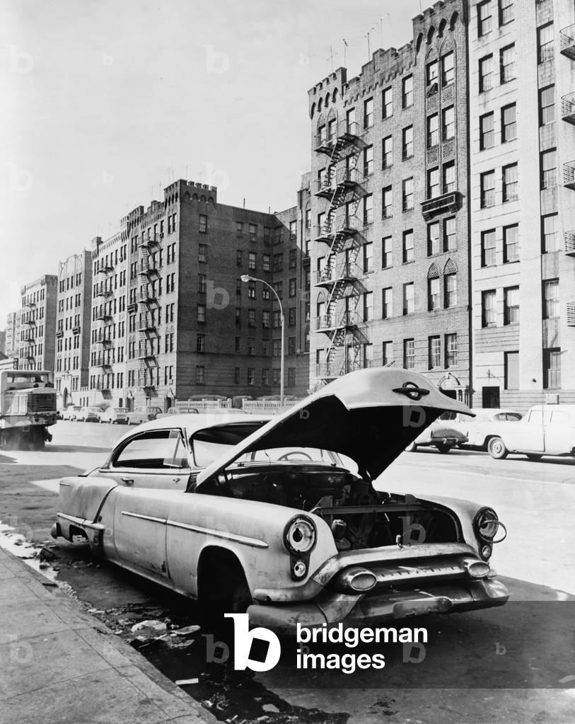 Bronx street in New York City. An abandoned automobile, a vacant lot and tenement apartments on Macombs Road. c. 1964