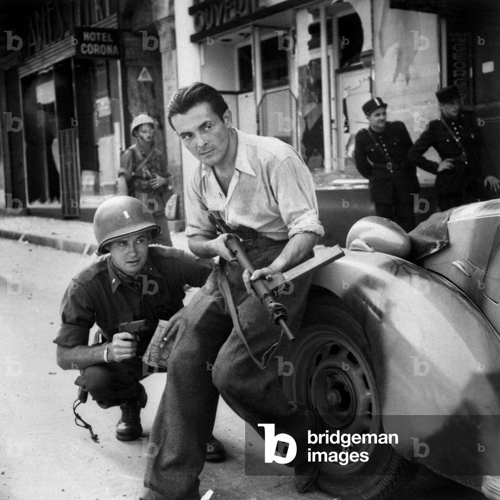 U.S. Army Lieutenant and French partisan crouch behind an auto in street fighting. Summer 1944. World War 2