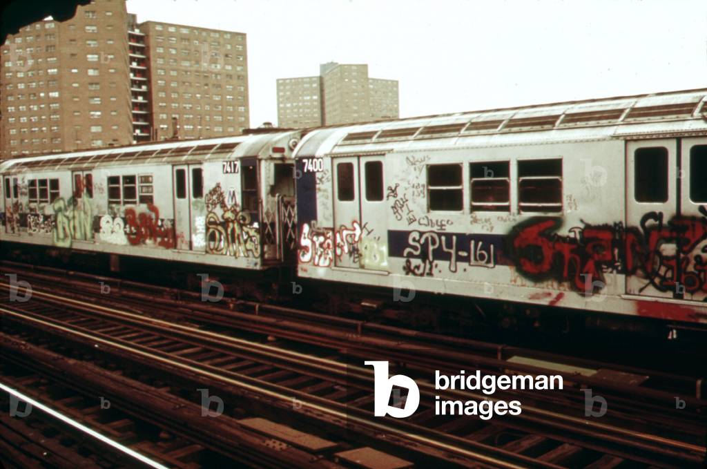 New York City Subway. A graffiti painted subway train with housing projects in the background. Outside of Manhattan many New York subways run on elevated tracks. May 1973