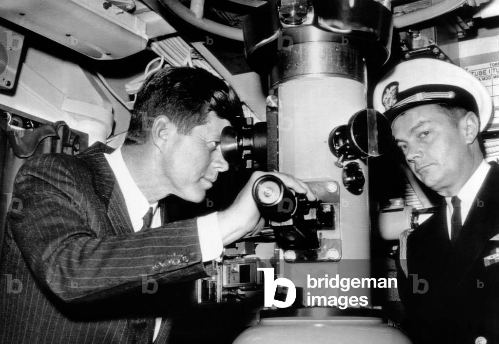 President John Kennedy looks through the periscope of the nuclear submarine USS Thomas A. Edison. At right is his Naval Aide, Capt. Tazewell Shepard