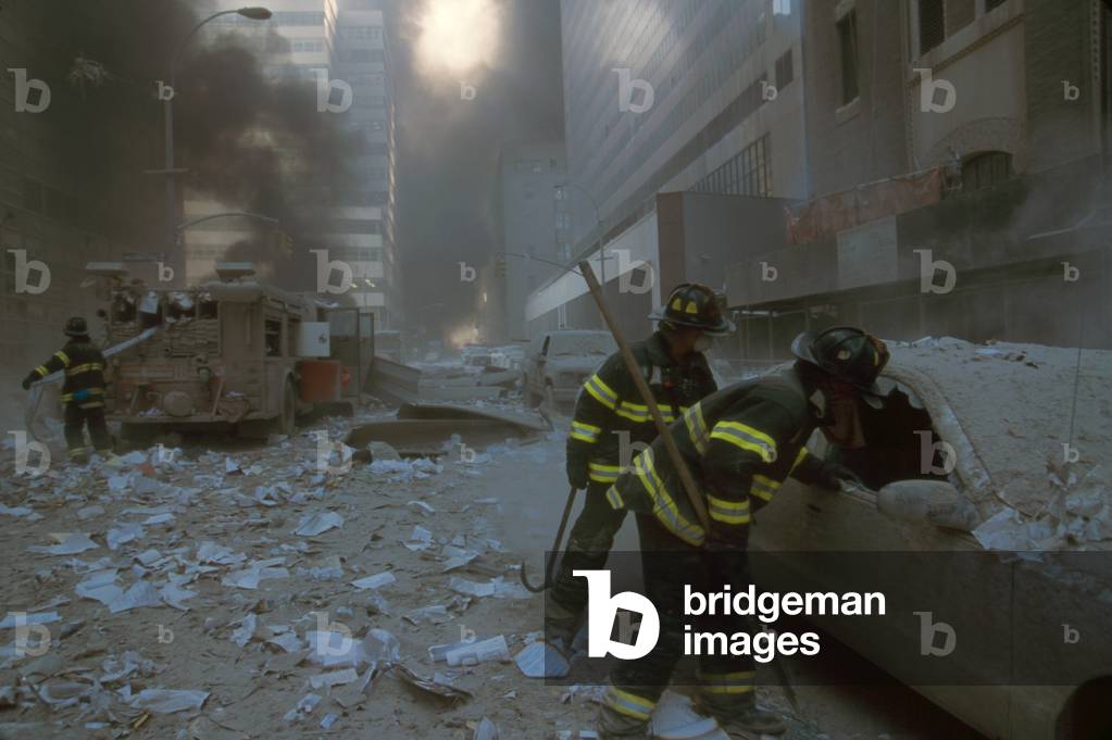 NYC firemen checking a car on Barclay Street after the 9-11 terrorist attack on World Trade Center. In the mid-ground fireman pulls a water hose from fire truck. View is toward West Broadway and includes WTC 7, on right, clad in red exterior masonry. WTC 7 collapsed at 5:20 PM on Sept. 11, 2001, due to fires ignited by debris from the North Tower (WTC 1) destruction earlier in the day
