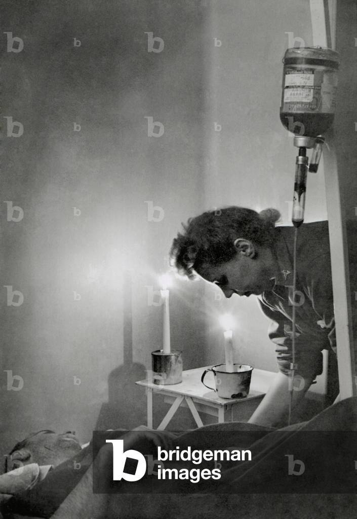 American Army nurse at candle lit bedside of wounded soldier in France during World War 2. He receives a transfusion at a front line hospital indicating a great lose of blood from his wound. Jan 23, 1945