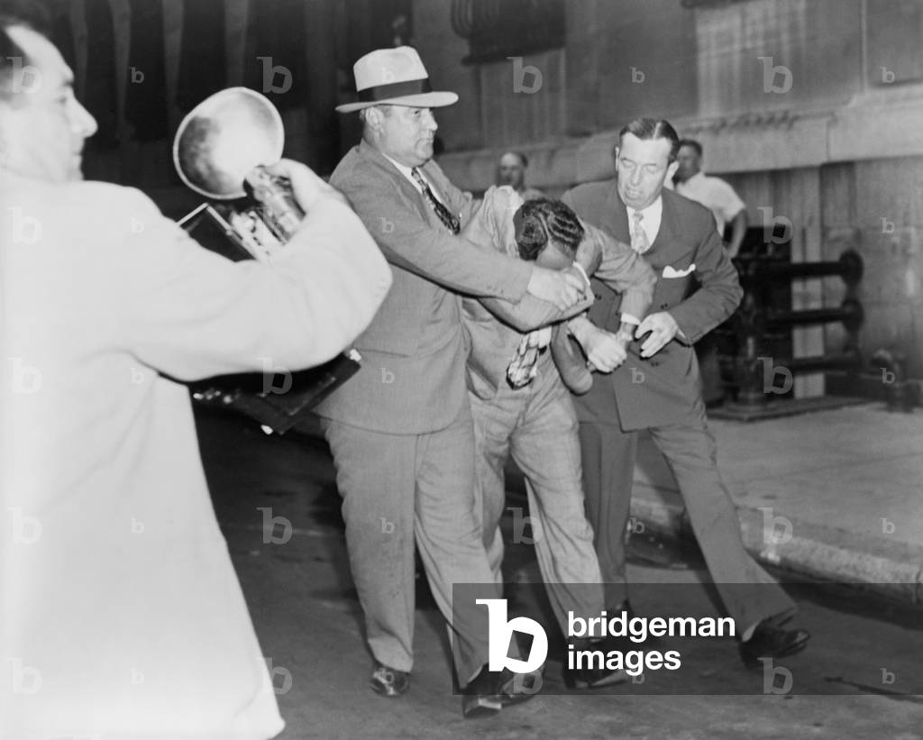 Two policemen forcibly escorting Vincent 'Jimmy Blue Eyes' Ado (1904-2001), after appearing in lineup; a news photographer (left) foreground captures the scene. August 1947