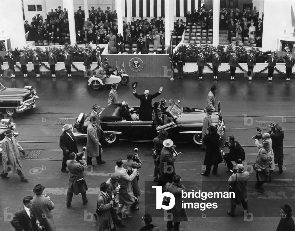 Eisenhower waves from an open car in the inaugural parade. Behind him is the White House Inaugural pavilion. Jan. 21, 1957.