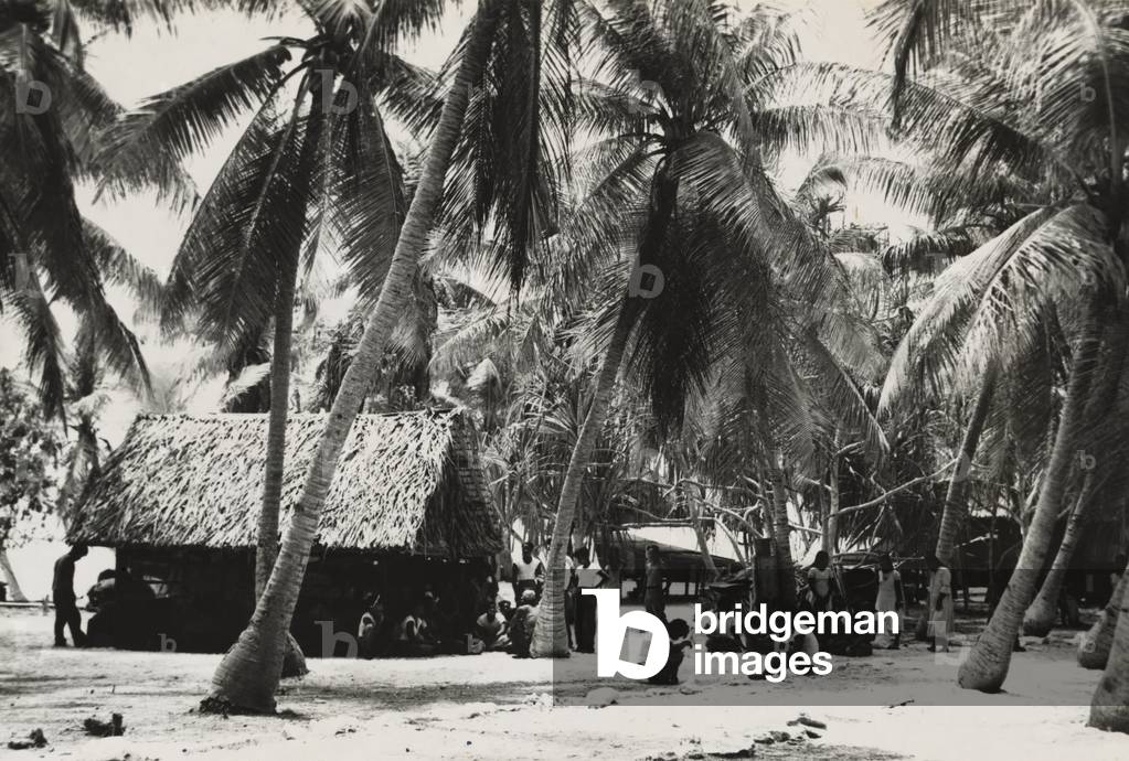 Bikini Islanders near thatched roof building shaded by palm trees, c. March 1946. 167 islanders from 40 families were required to move to Rongerik Atoll by U.S. Military before the Operations Crossroads atomic bomb tests of July 1946