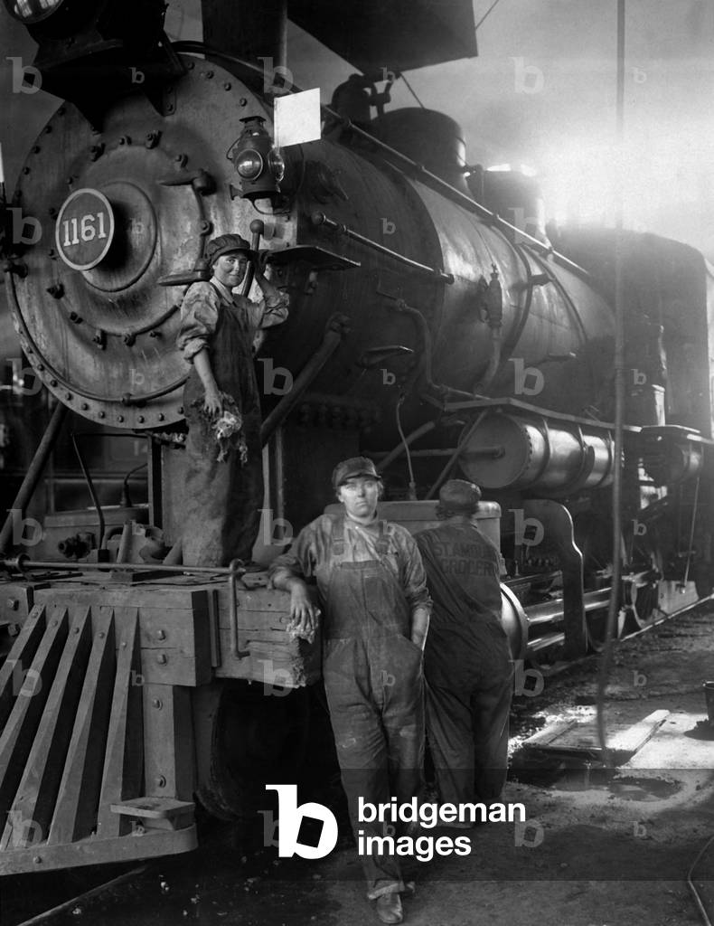 Women taking the place of men on Great Northern Railway at Great Falls, Montana. They are wiper's, doing a dirty, entry level job, cleaning surfaces of the train engines and machinery. World War I. c. 1918