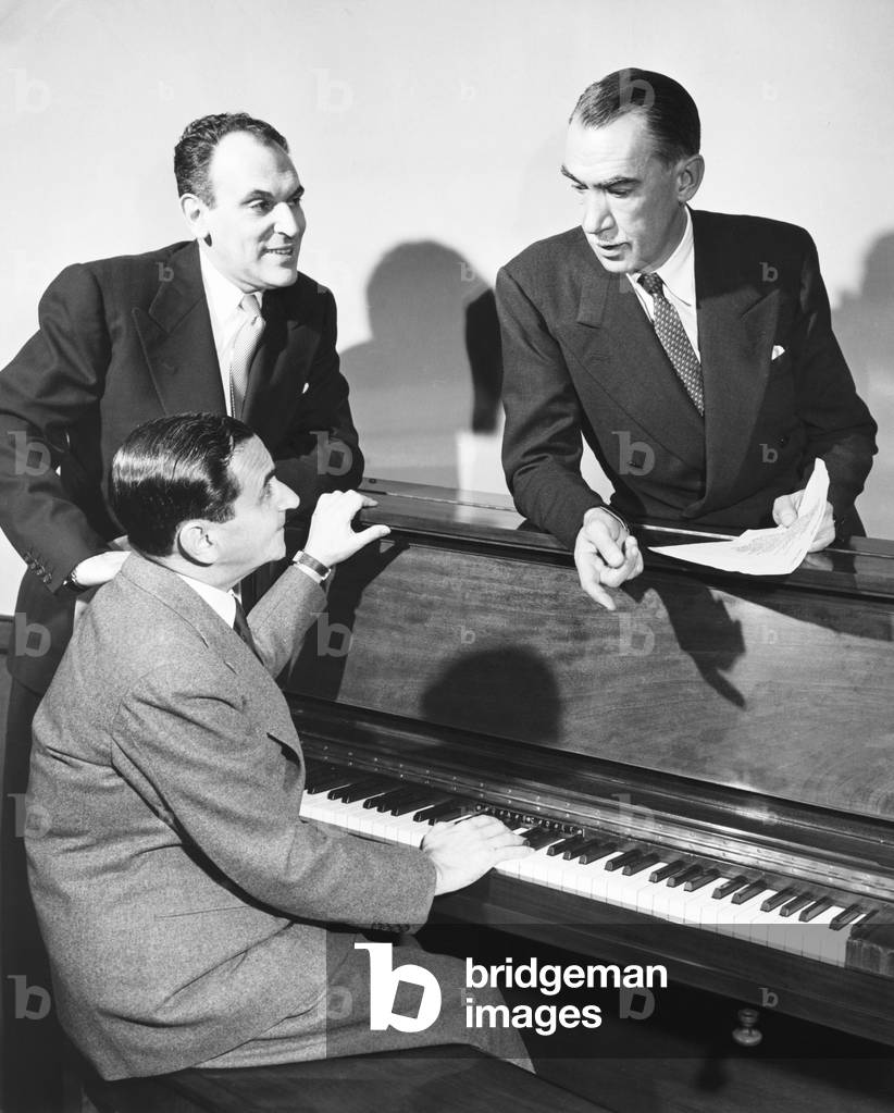 Moss Hart (left), Robert E. Sherwood, and Irving Berlin at the piano. They are collaborating on a new musical, 'Miss Liberty' based on a book by Robert E. Sherwood with music and lyrics by Irving Berlin. The score includes the song 'Give Me Your Tired, Your Poor,' a musical setting of the poem by Emma Lazarus. March 1949. Photo by Hugelmeyer
